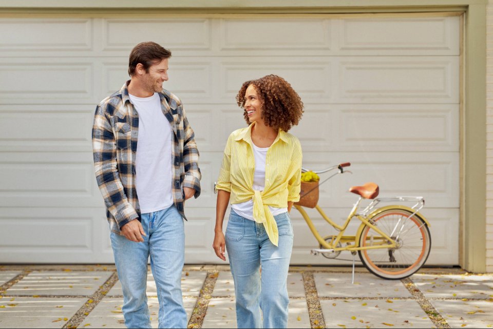 Two people walking side by side on a paved driveway in casual clothing, with a yellow bicycle parked in front of a garage behind them.