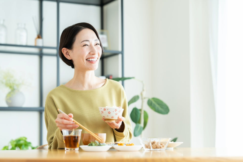 A smiling woman with short dark hair sits at a wooden table in a bright kitchen, holding a small floral-patterned bowl and chopsticks. She wears a muted olive-green top, with dishes and a glass of tea in front of her.