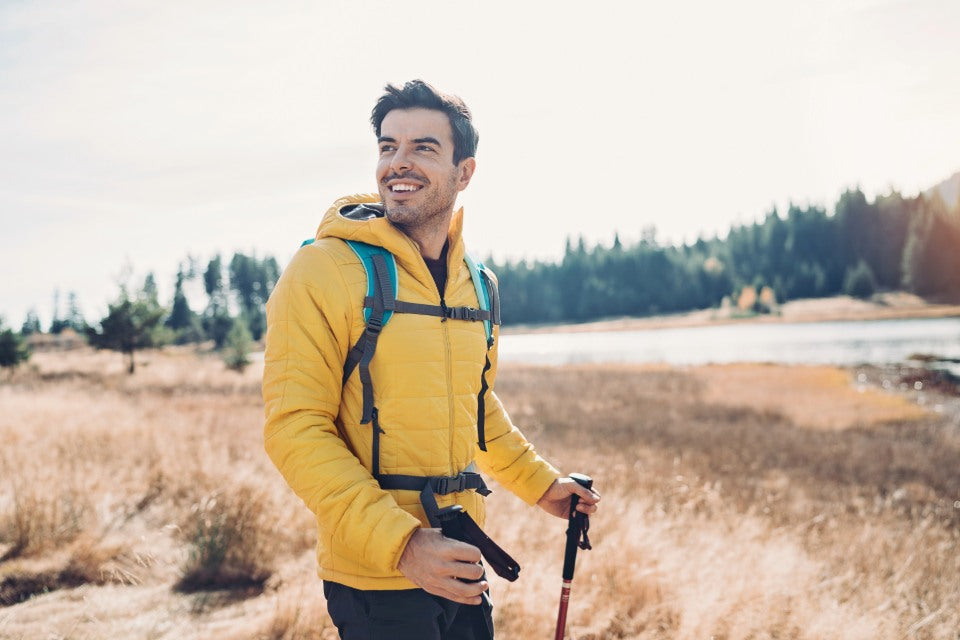 Man in yellow jacket with backpack and hiking poles standing in a field with trees and water in the background