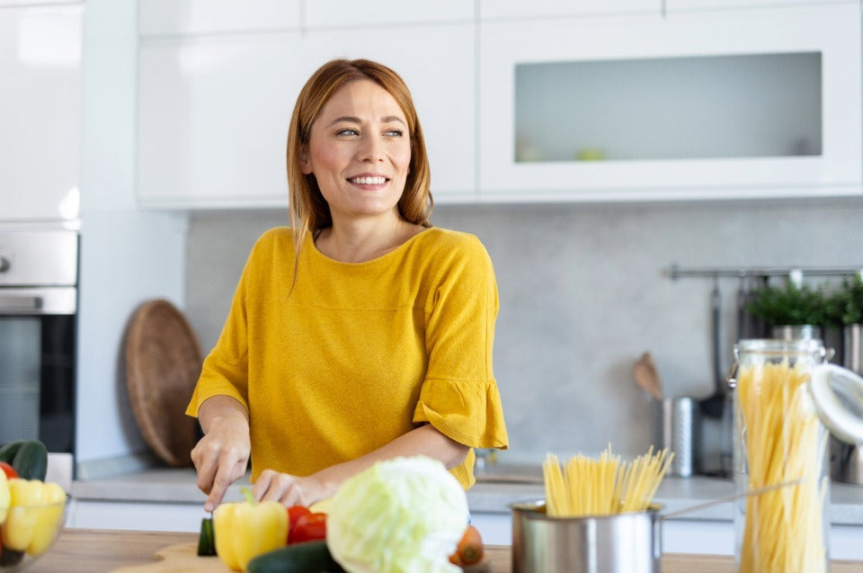 Person in a yellow top chopping fresh vegetables on a kitchen counter with pasta nearby.
