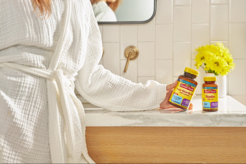 Person in a bathroom holding Nature Made Hair, Skin & Nails supplement next to a Biotin bottle on a bathroom counter