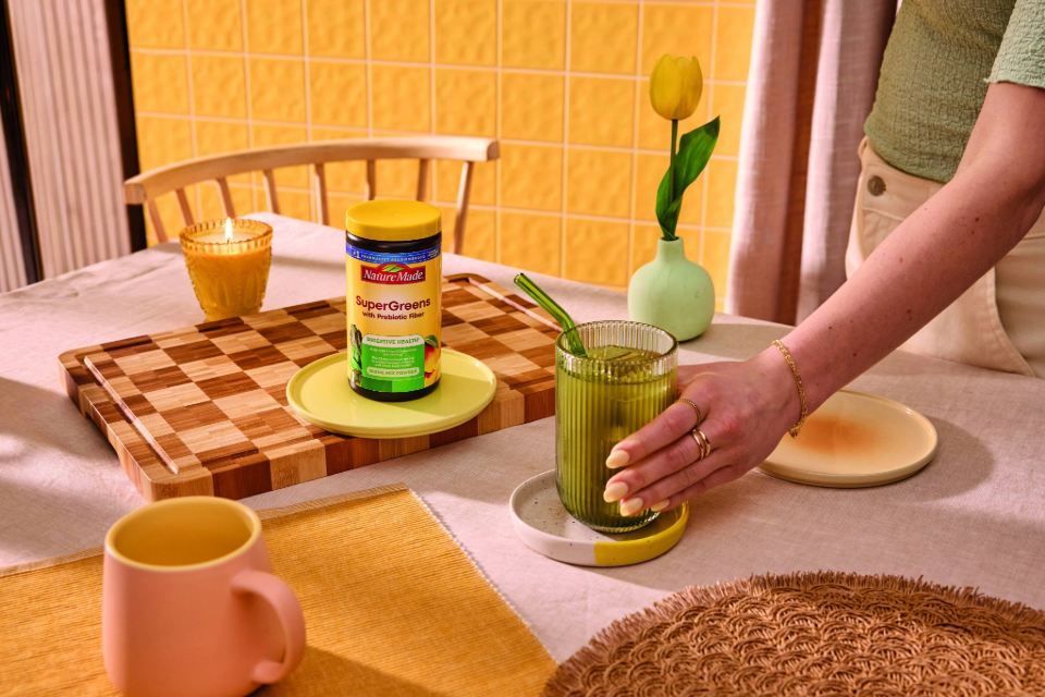 Nature Made SuperGreens with Prebiotic Fiber Mix and a glass of mixed greens on a dining table.