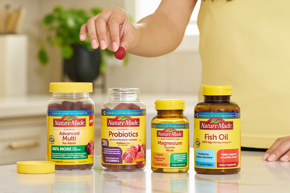 Hand picking a gummy from Nature Made supplement bottles on a kitchen counter, featuring Advanced Multi, Probiotics, Magnesium, and Fish Oil.