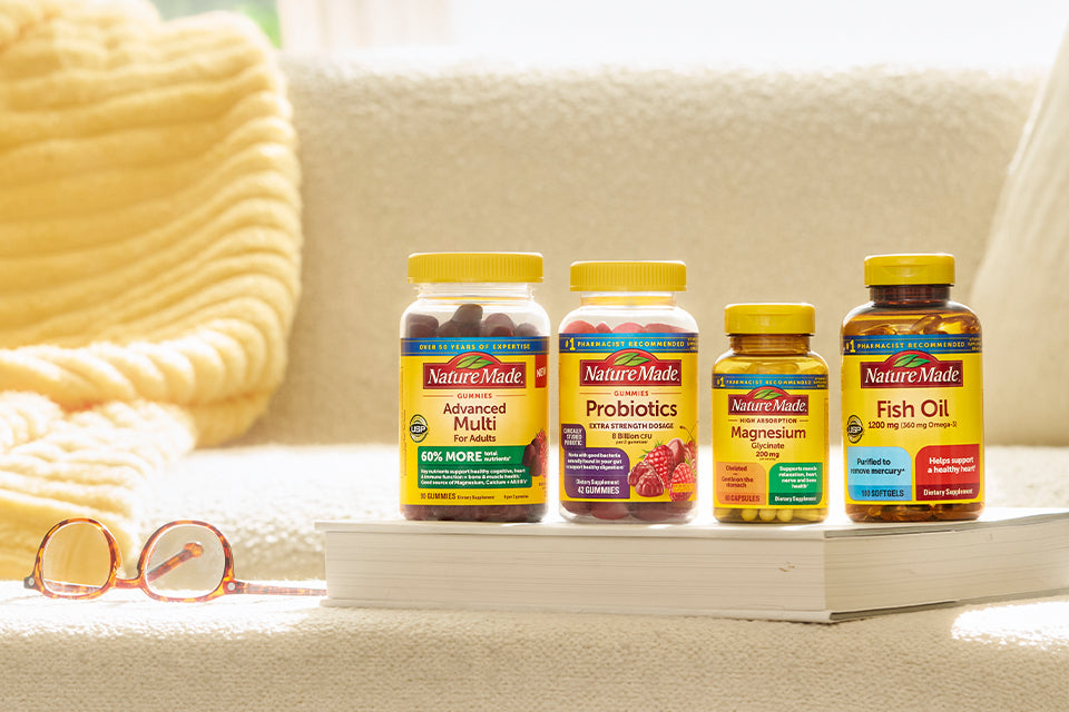 Nature Made vitamins and supplements on a cozy couch with a yellow blanket and glasses, featuring Advanced Multi, Probiotics, Magnesium, and Fish Oil bottles.