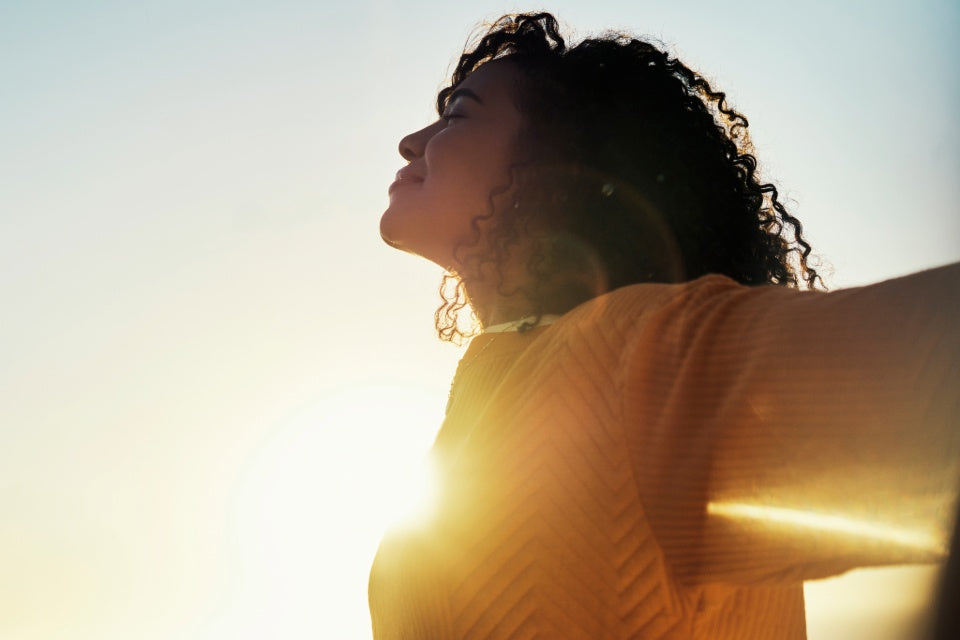 Person standing outdoors with arms outstretched in sunlight, wearing a yellow sweater.