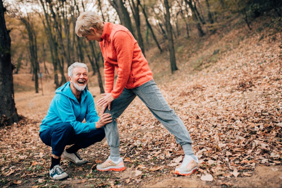 Two older adults exercising outdoors in a wooded park, with one assisting the other during a leg stretch on a leaf-covered trail.