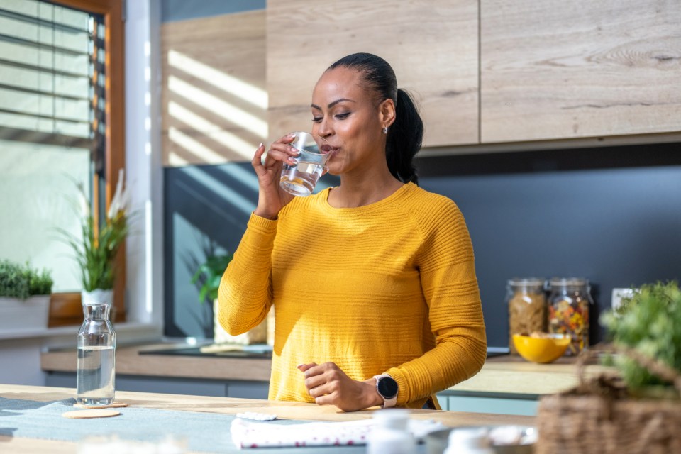Person in a yellow sweater drinking a glass of water in a modern kitchen.