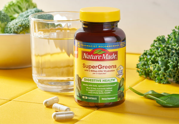 Nature Made SuperGreens capsules supplement bottle on a yellow surface with broccoli and a glass of water in the background.
