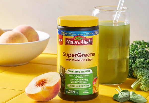 Nature Made SuperGreens drink mix powder with a glass of green juice, peaches, and broccoli on a yellow surface.