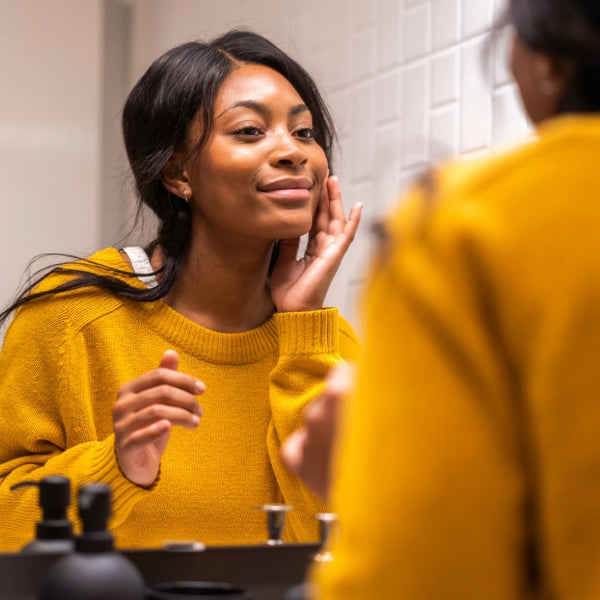 A woman in a yellow knit sweater looks into a bathroom mirror while touching her face. 